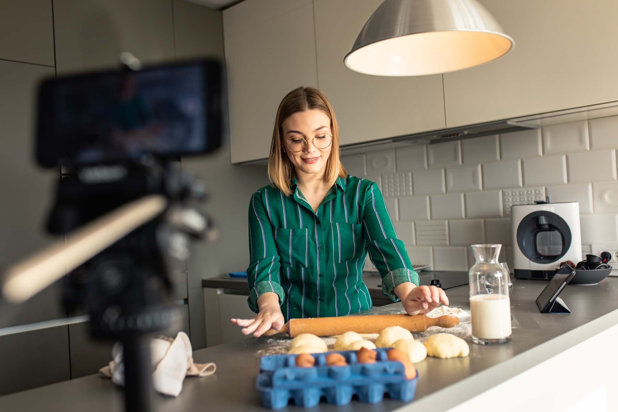 Young woman recording video blog with smart phone at home in kitchen making croissants.