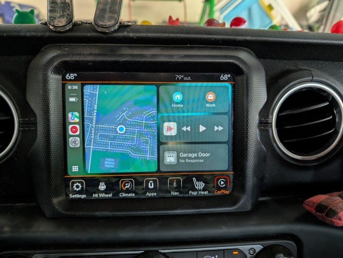 A close-up of a car's infotainment screen displaying a map and various apps, including climate controls and connectivity options. The dashboard has a textured finish with visible air vents and a plaid-patterned cloth.