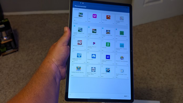 A person holding a Lenovo tablet displaying a screen filled with recommended apps and games, set against a neutral background.