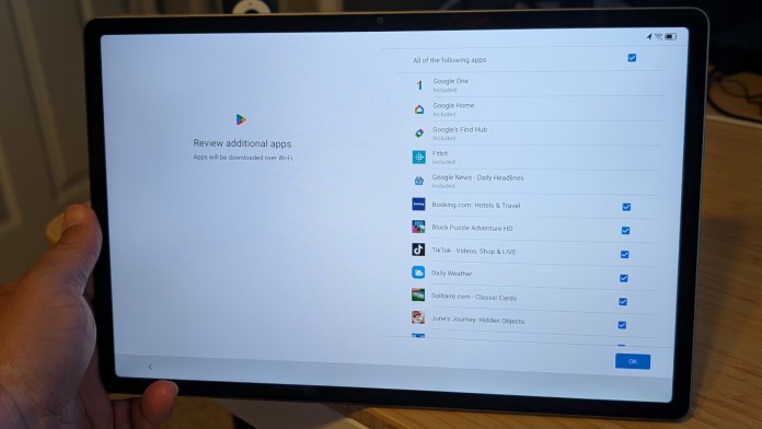 A hand holding a Lenovo tablet displaying a screen for reviewing additional apps to be downloaded over Wi-Fi, with several app options listed.