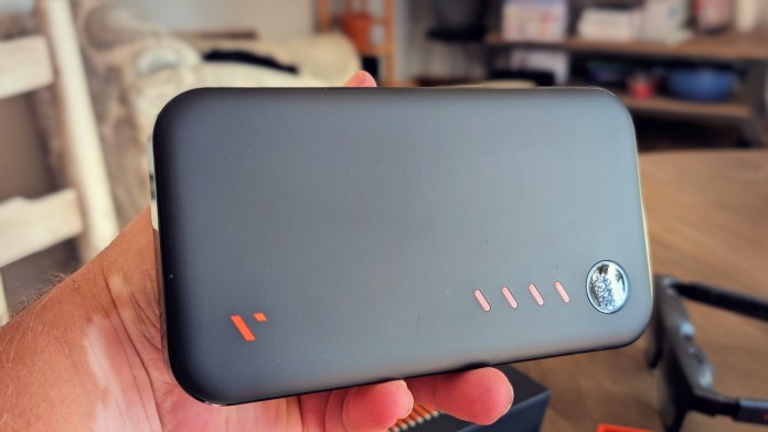 A person holding a compact black power bank with an orange logo and indicator lights, set against a casual indoor background.