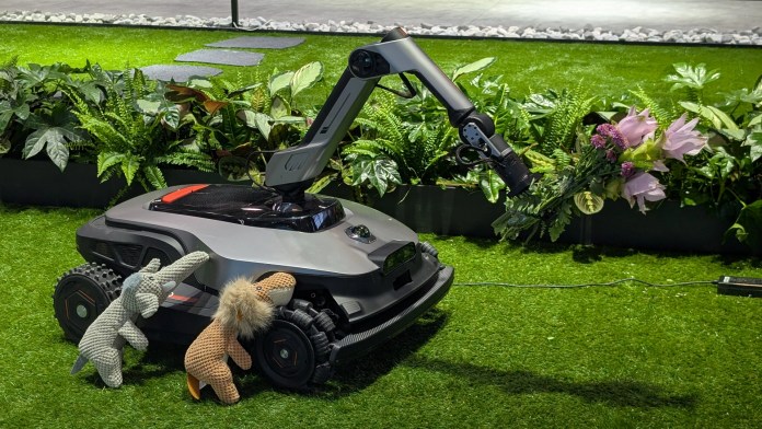 A robotic lawn mower with articulated robotic arm on a patch of artificial grass, decorated with plush animal toys, alongside various indoor plants.