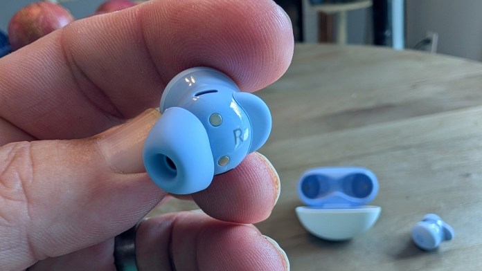 Close-up of a hand holding a blue Pixel Buds 2a earbud, showcasing its sleek design and silicone tip with a charging case in the background.
