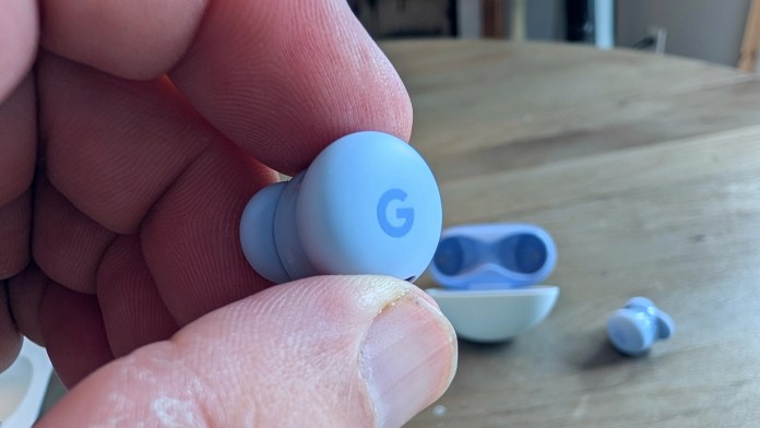 A close-up shot of a person's hand holding a blue Pixel Buds 2a earbud with a visible 'G' logo, with another earbud and the charging case in the background.