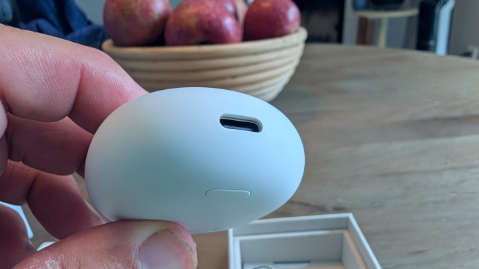 A close-up image of a hand holding the case of Google Pixel Buds 2a, showing the USB-C charging port. In the background, a bowl of red apples is visible on a wooden table.