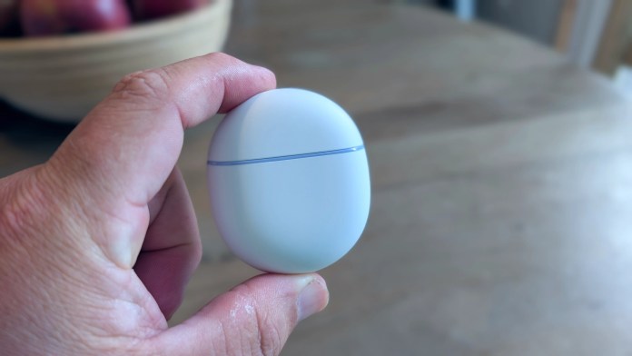 A person holding a white charging case for Google Pixel Buds 2a, with a wooden table and a bowl of apples in the background.