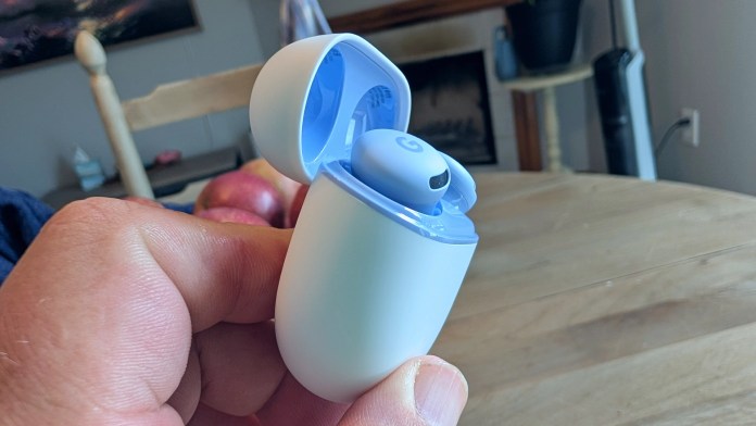 A person holding a Google Pixel Buds 2a case open, revealing the earbuds inside. The background features a table with apples and a cozy home setting.
