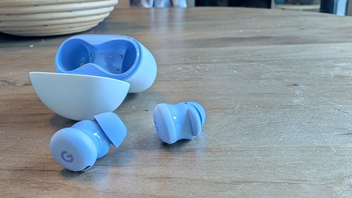 The image shows Google Pixel Buds 2a in a light blue color, with a matching pebble-shaped charging case partially open. Two earbuds are placed on a wooden surface next to the case.