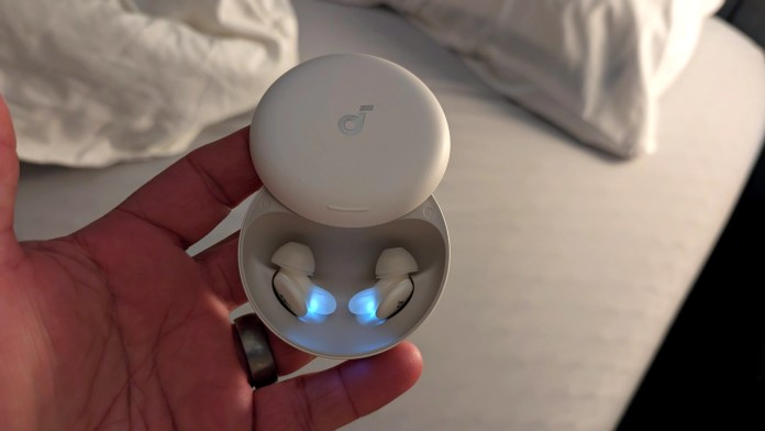 A person holding a charging case for Soundcore Sleep A30 earbuds, with the earbuds illuminated inside, against a soft bedding background.