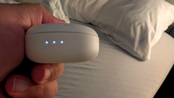 A person holding a charging case with three blue LED lights, positioned above a bed with white pillows.