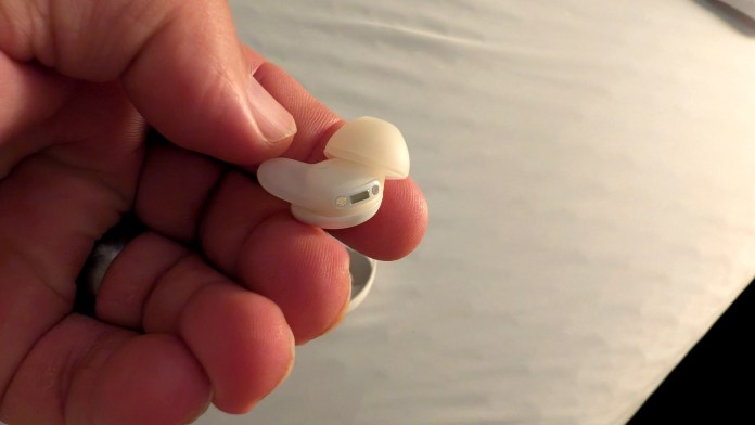 A person holding a white wireless sleep earbud with a soft silicone tip, against a blurred background of a bed.