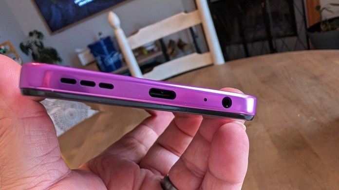 A close-up image of a hand holding a purple smartphone, showcasing the bottom edge with audio ports and speaker grilles.