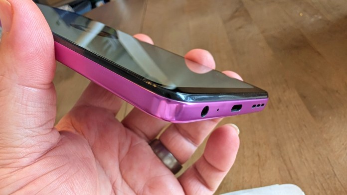 A person holding a smartphone with a pink metallic edge, showing the bottom view with charging port, headphone jack, and speaker grille.