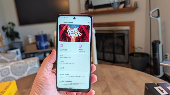 A person holding a smartphone displaying the 'About phone' section with device information like model name, storage, RAM, camera specifications, and display resolution in a cozy indoor setting.