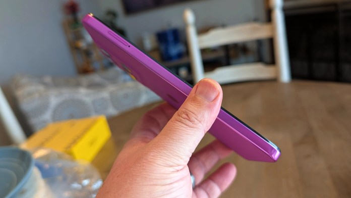 A person holding a slim purple smartphone in their hand, viewed from the side, with a blurred background of a living area.