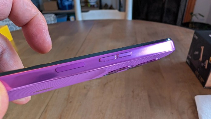 A close-up view of a purple smartphone held at an angle, showcasing its side buttons and a textured surface, with a blurred background of a wooden table and other objects.
